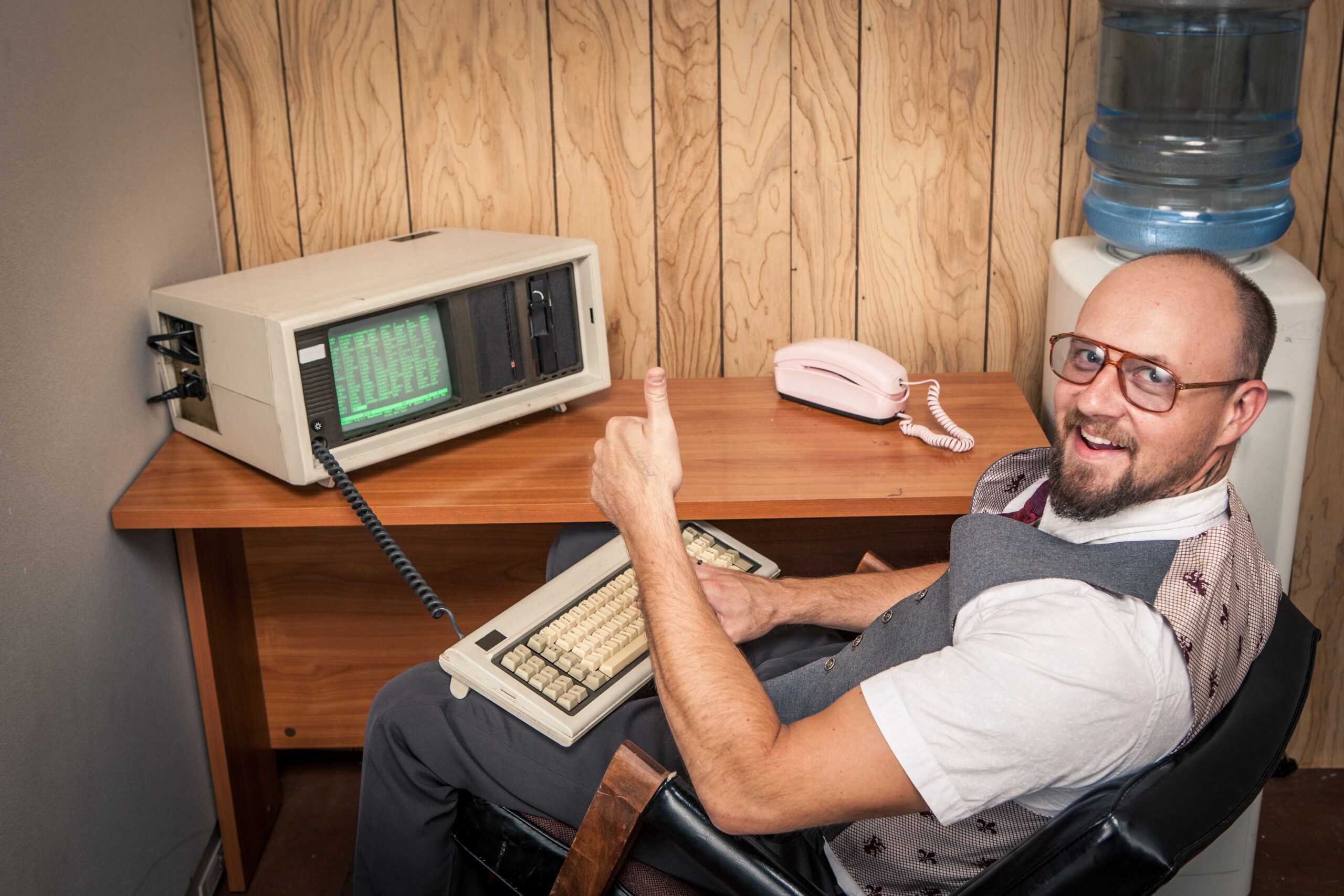 Happy office worker sitting, typing, working, and selling big at 1970's or 1980's monochrome computer.  Retro office interior scene.  Wood Paneling and a water cooler in the background. Old retro vintage technology